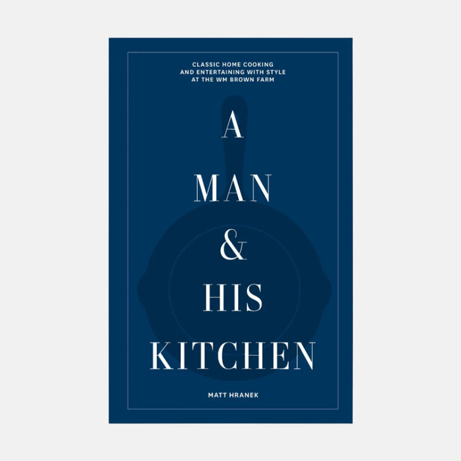 A Man & His Kitchen" hardcover cookbook with dark blue cover, white text, and subtle cast iron pan graphic.