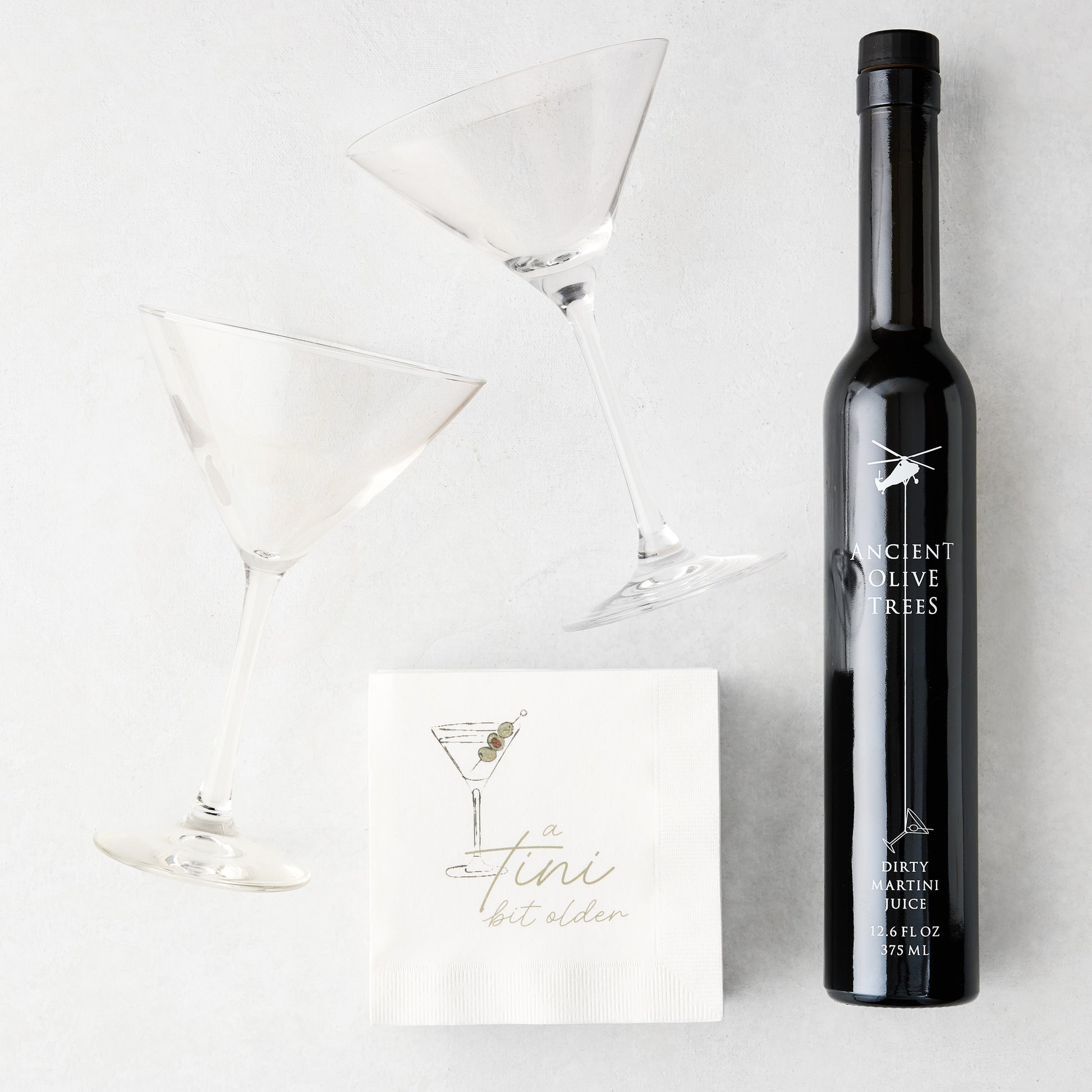 Two clear martini glasses, a black bottle of Ancient Olive Trees Dirty Martini Juice, and white "a tini bit older" cocktail napkins.