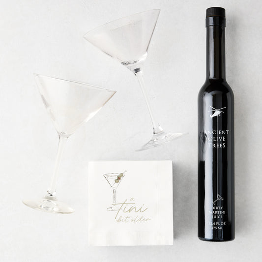 Two clear martini glasses, a black bottle of Ancient Olive Trees Dirty Martini Juice, and white "a tini bit older" cocktail napkins.