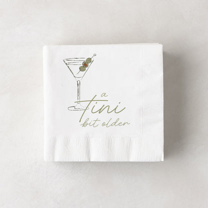 White 3-ply paper cocktail napkins with a martini glass and "a tini bit older" in olive green.