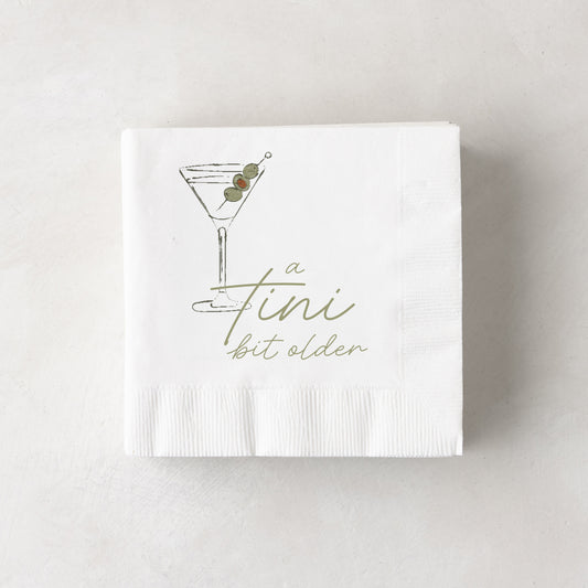 White 3-ply paper cocktail napkins with a martini glass and "a tini bit older" in olive green.