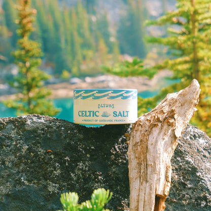 Altura Celtic Salt tin, light blue and cream, on dark rock with driftwood and green trees.
