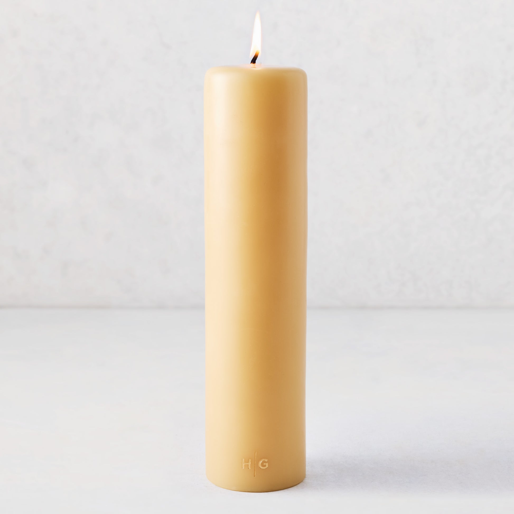 3x12 inch amber unscented paraffin pillar candle with a lit wick and embossed gold HG logo on a light background.