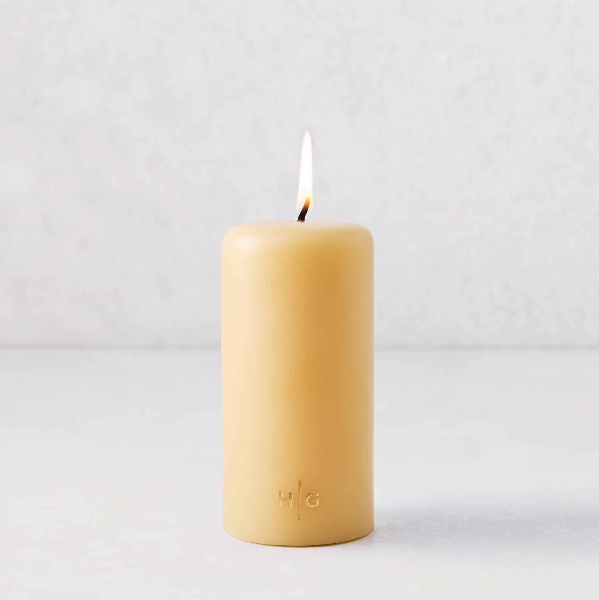 3x6 inch amber pillar candle with a lit cotton wick and gold embossed HG logo on a white background.