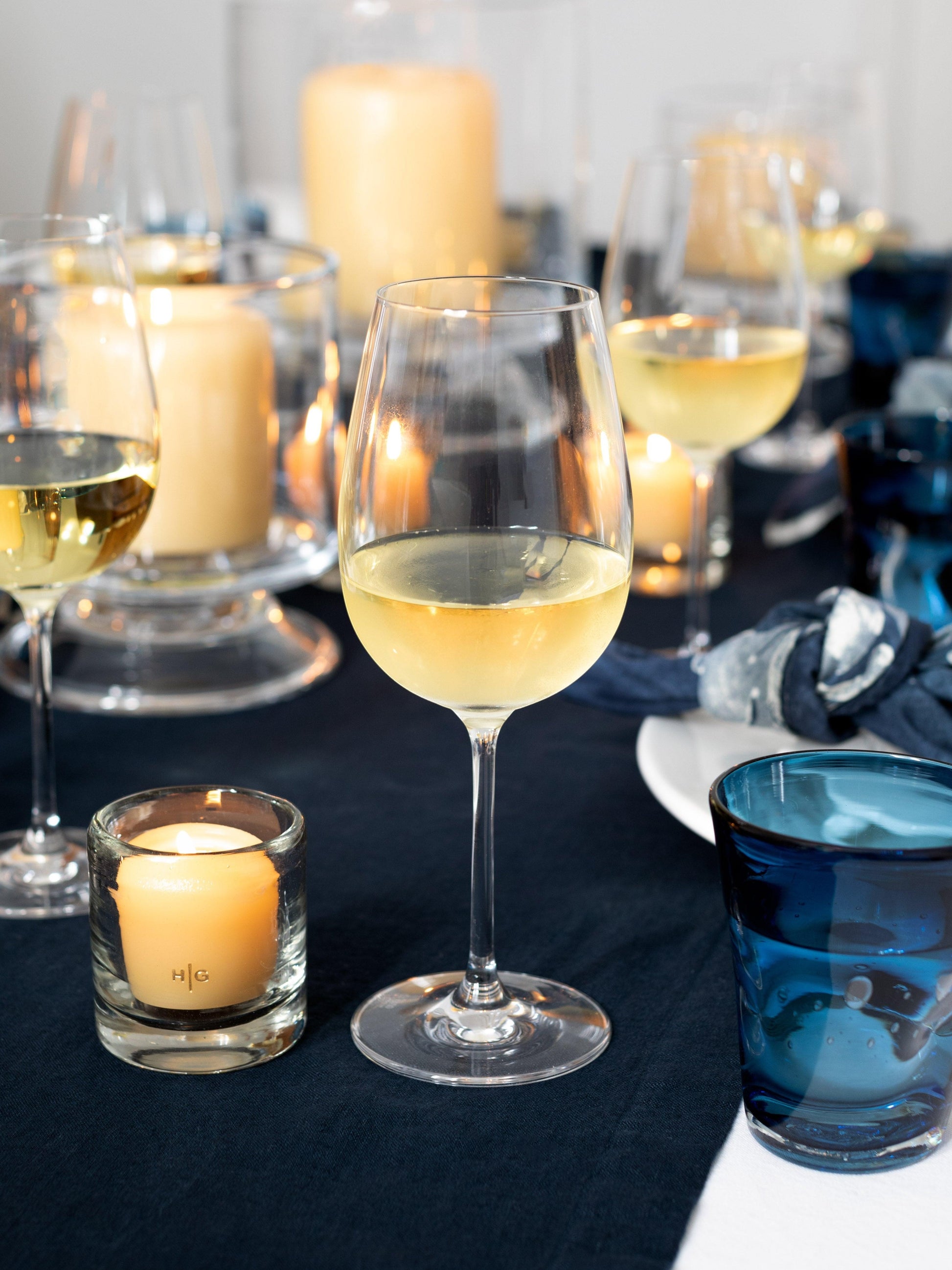 HG Amber Unscented 2" Votive Candle in a glass holder, with a gold HG logo, on a dark blue tablecloth.