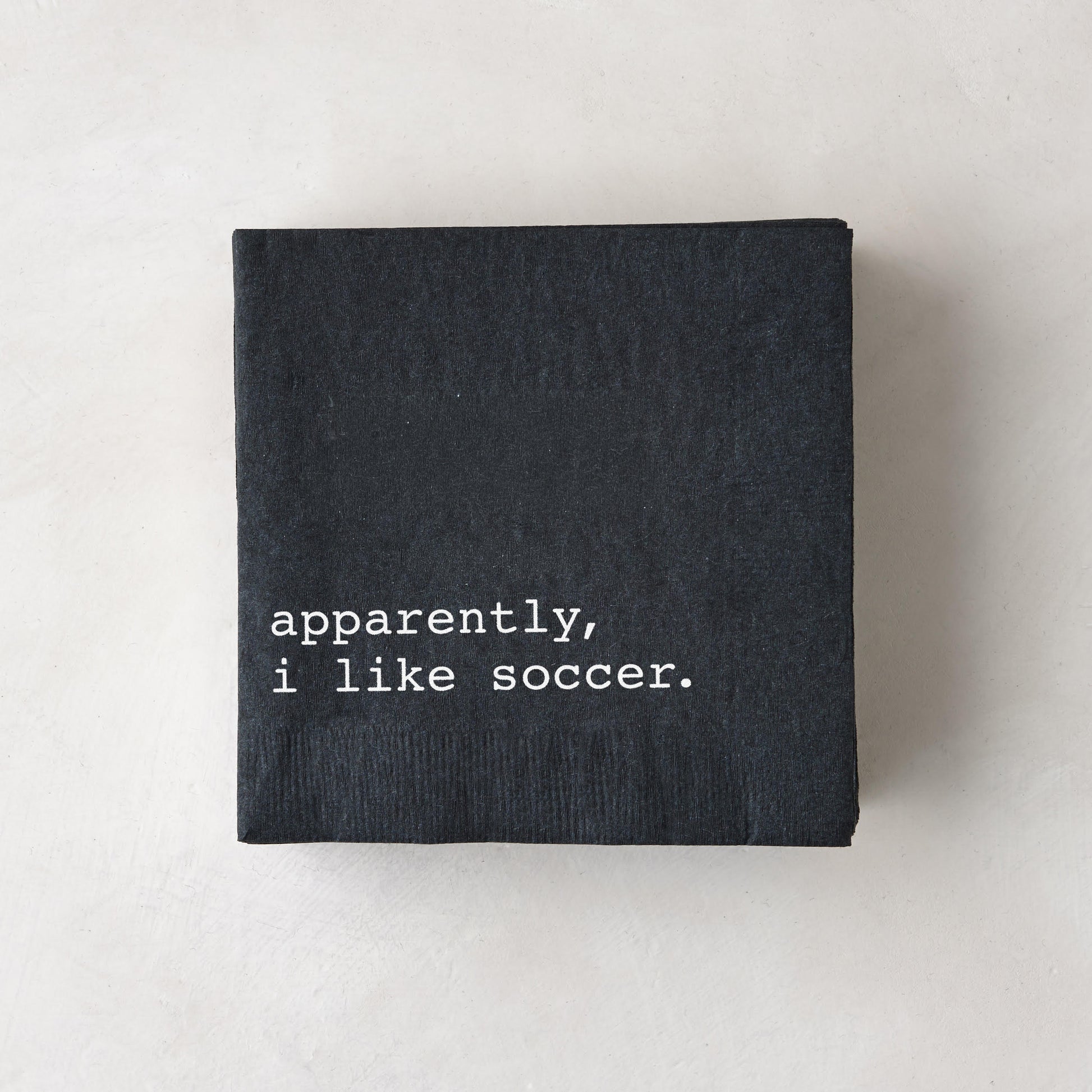 Black 5.5" square paper cocktail napkin with "apparently, i like soccer." in white text.