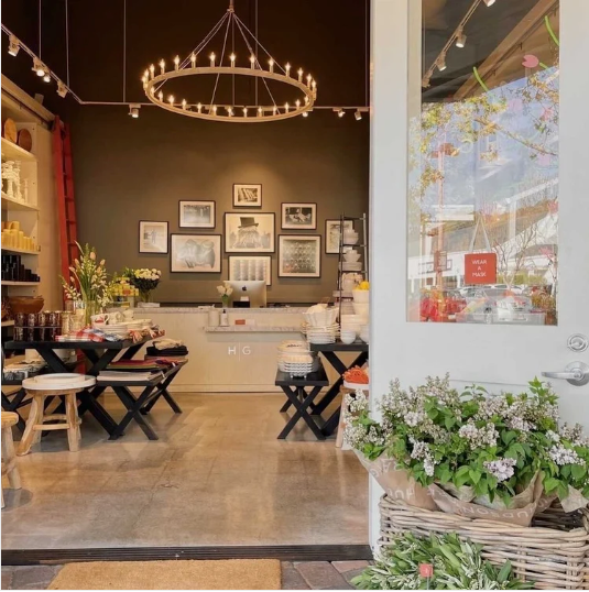 Boutique interior with dark walls, framed art, a large chandelier, and tables displaying home goods.