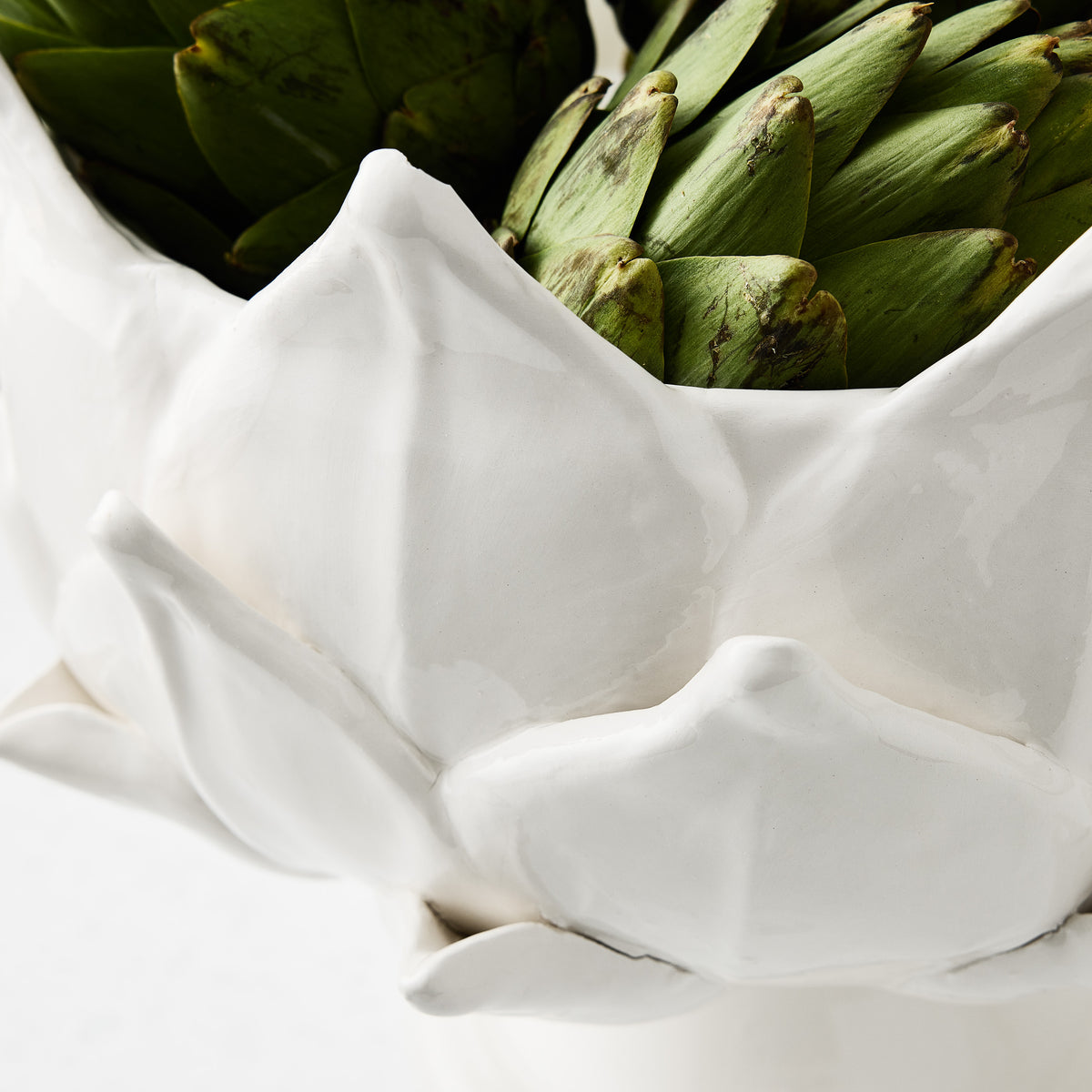 White ceramic footed bowl shaped like artichoke leaves, holding fresh green artichokes, 14-inch diameter.