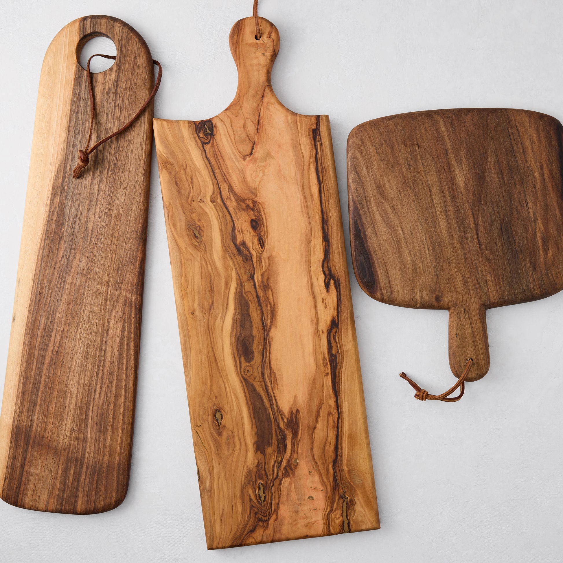 Three unique wood serving boards: long walnut, olive wood with rich grain, and a dark rectangular board, all with leather cords.