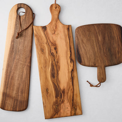 Three unique wood serving boards: long walnut, olive wood with rich grain, and a dark rectangular board, all with leather cords.