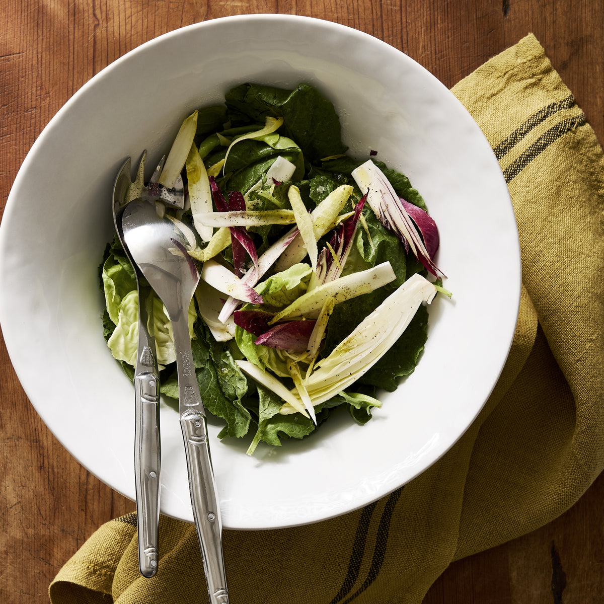 Large, creamy white stoneware serving bowl filled with salad and silver serving utensils on a wooden table.