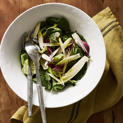 Large, creamy white stoneware serving bowl filled with salad and silver serving utensils on a wooden table.