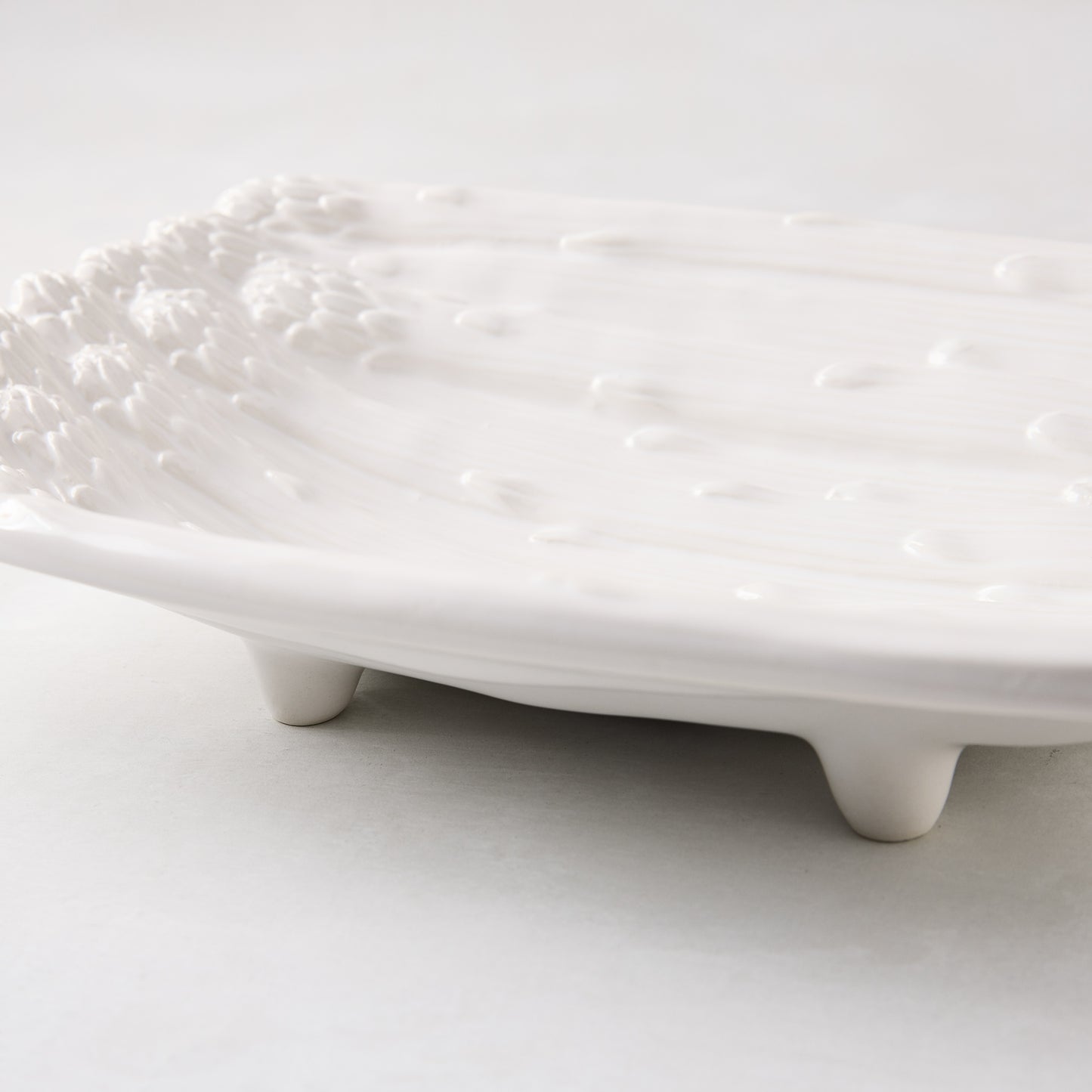 White ceramic serving dish with raised asparagus design and small feet.