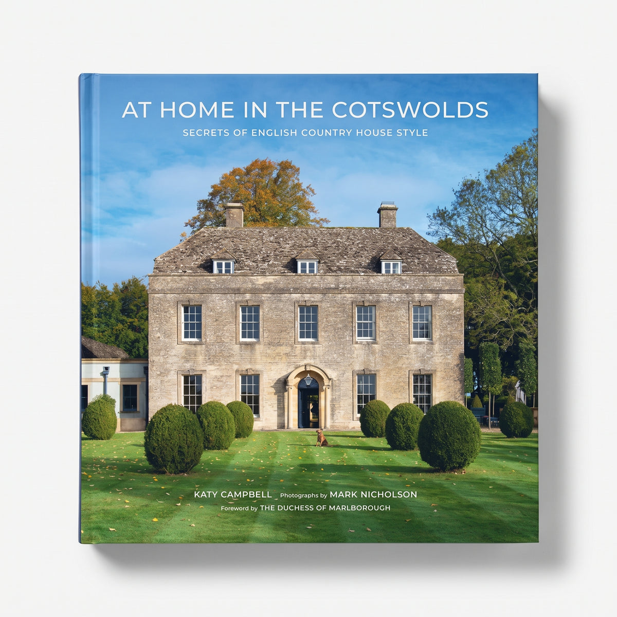 Hardcover book titled At Home in the Cotswolds featuring a stone English country manor and manicured garden on cover.