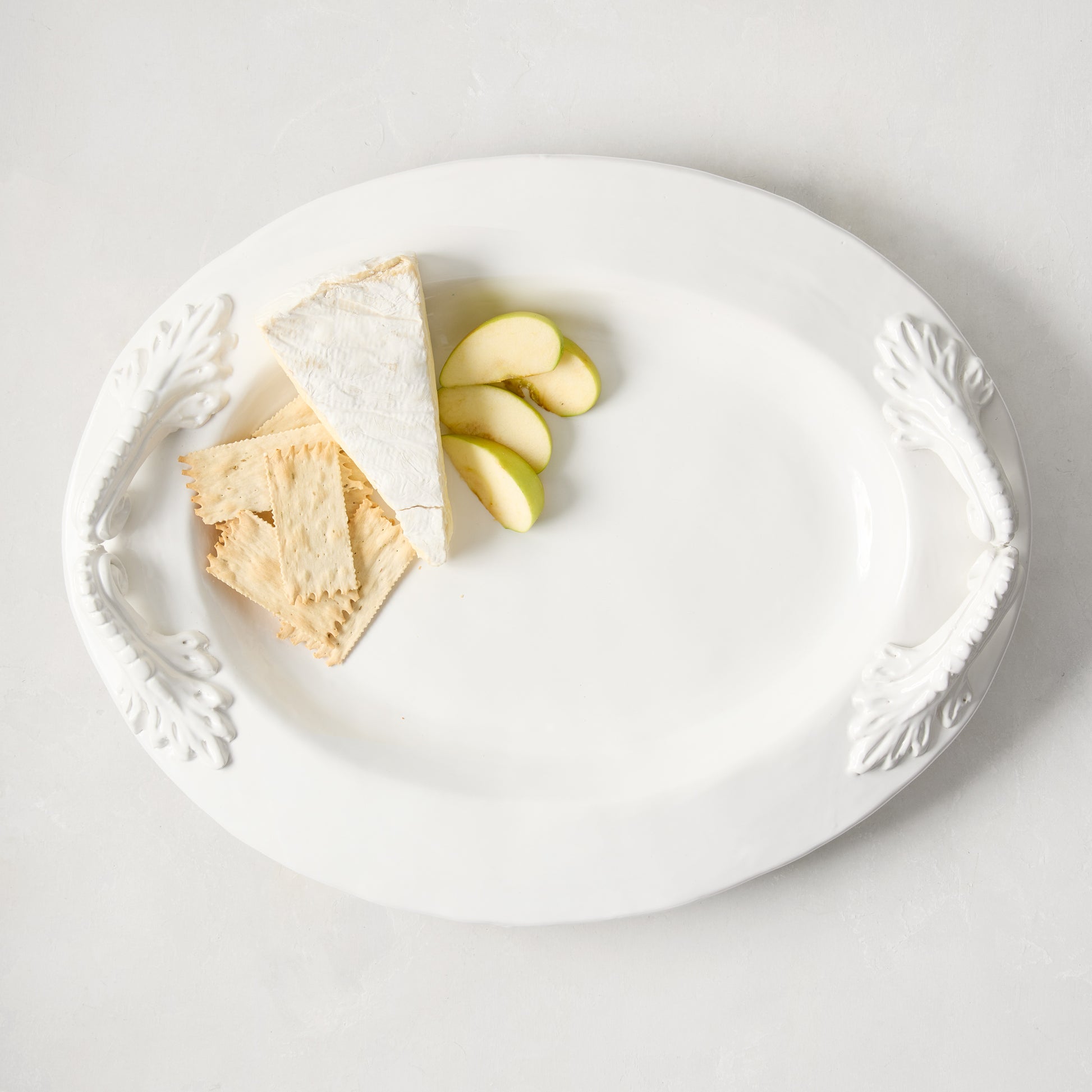 White ceramic oval serving platter with ornate handles, holding brie, crackers, and apple slices.