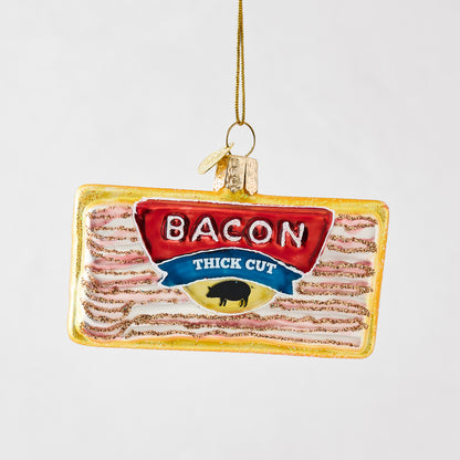 Hand-painted glass bacon ornament with glitter accents, gold hanging string, and "Bacon Thick Cut" label design.
