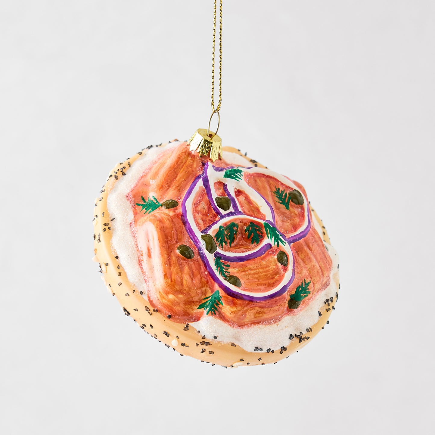 Hand-painted glass bagel and lox Christmas ornament with cream cheese, red onion, capers, and poppy seed details.