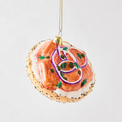 Hand-painted glass bagel and lox Christmas ornament with cream cheese, red onion, capers, and poppy seed details.