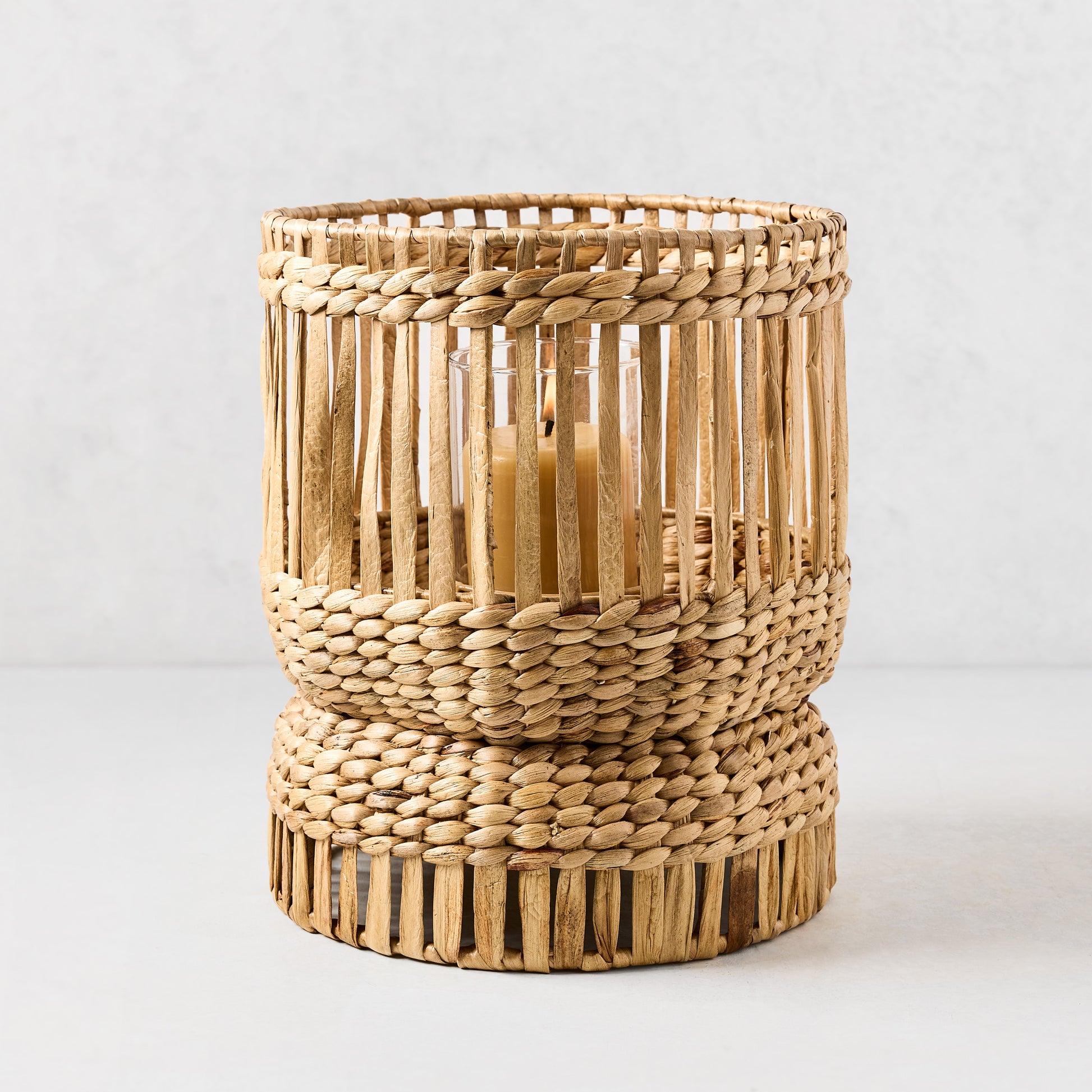 Bali Large Woven Hyacinth Hurricane with a lit candle, natural brown, cylindrical, artisan design.
