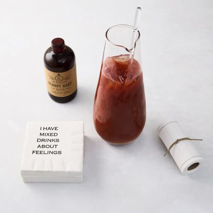 Clear glass cocktail jug with stirrer, filled with a red drink, next to a Bloody Mary mixer bottle and napkins.