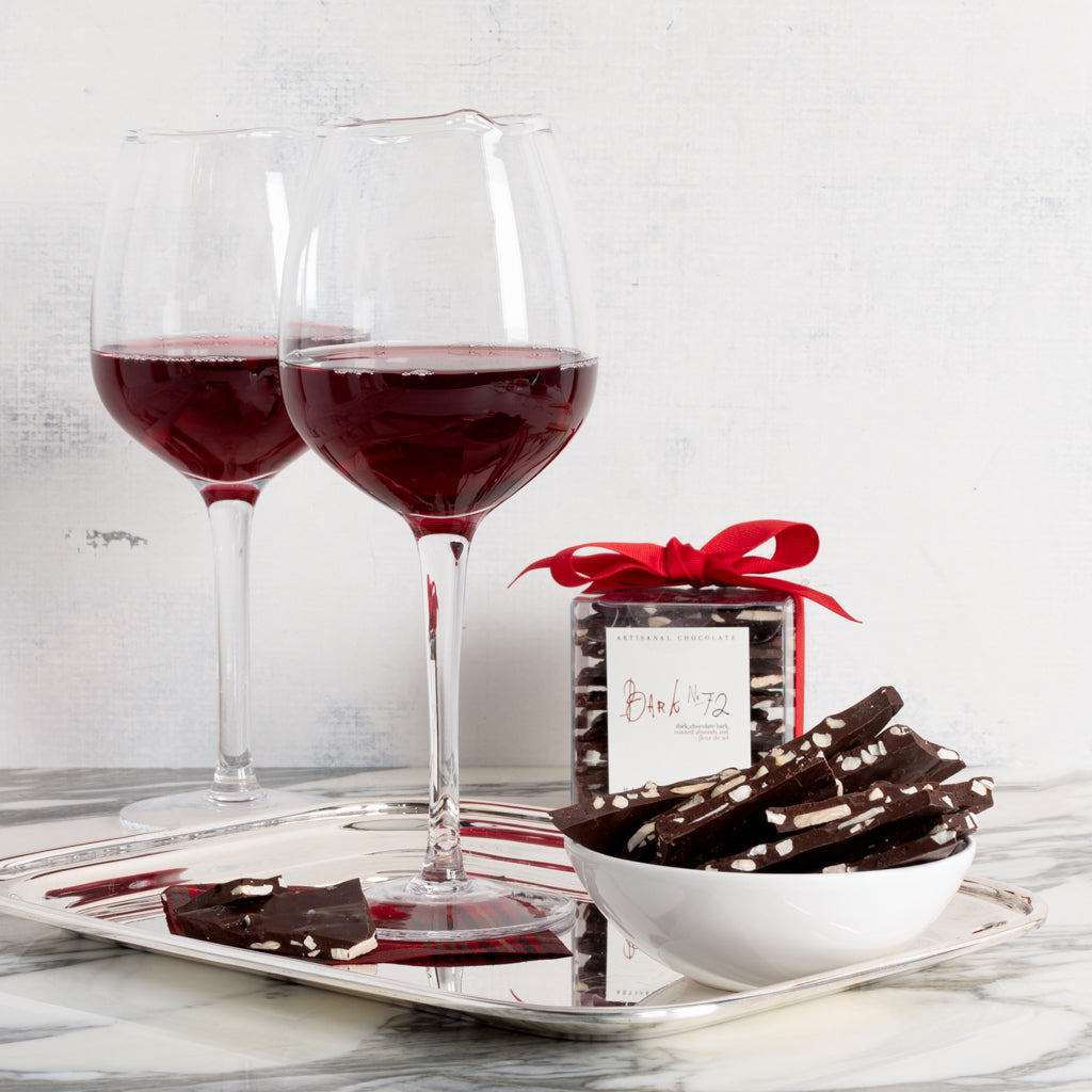 Bark 72 Dark Chocolate Almond Bark in a clear box with red ribbon, a white bowl of bark pieces, and two red wine glasses on a silver tray.