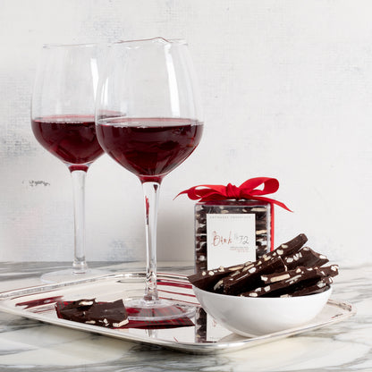 Bark 72 Dark Chocolate Almond Bark in a clear box with red ribbon, a white bowl of bark pieces, and two red wine glasses on a silver tray.