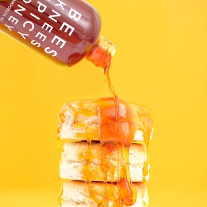 Bees Knees Spicy Honey in a clear bottle pouring amber honey over a stack of biscuits on a yellow background.