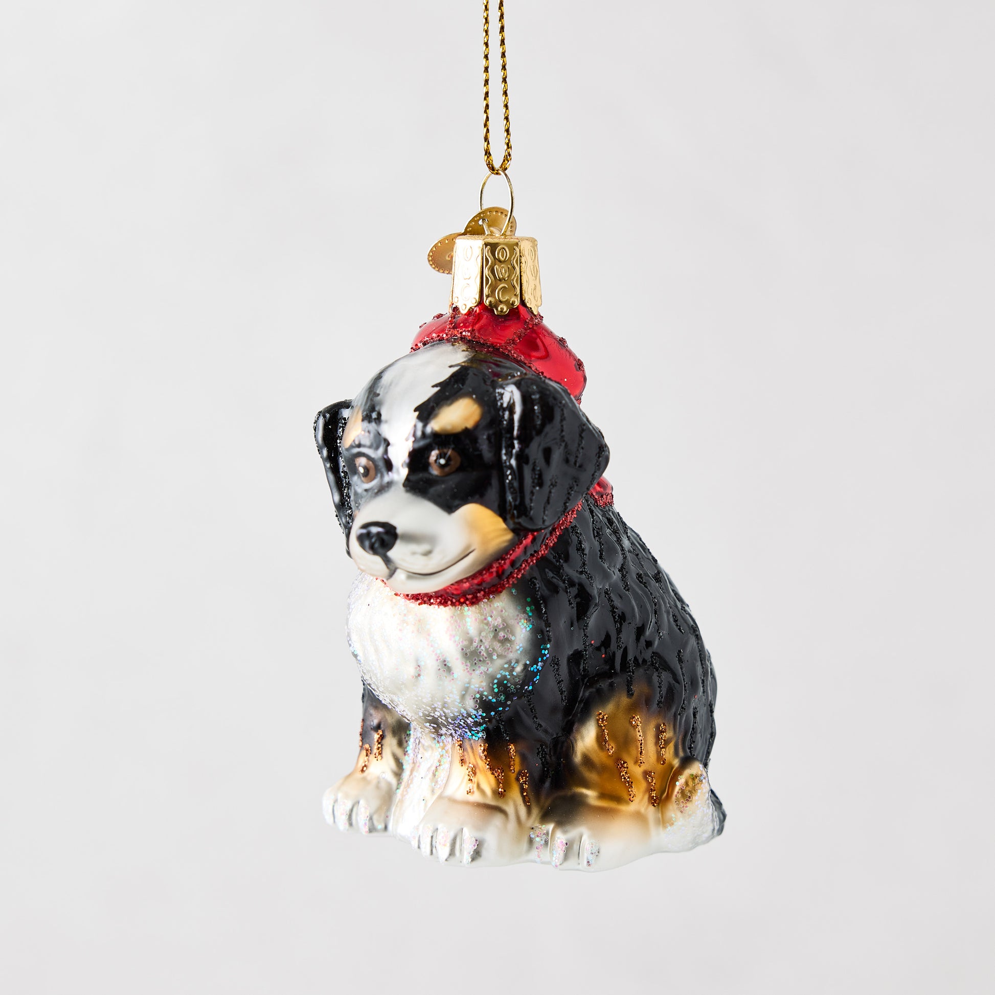Hand-painted glass Bernadoodle ornament wearing a red hat with glitter accents and a gold hanging cord.