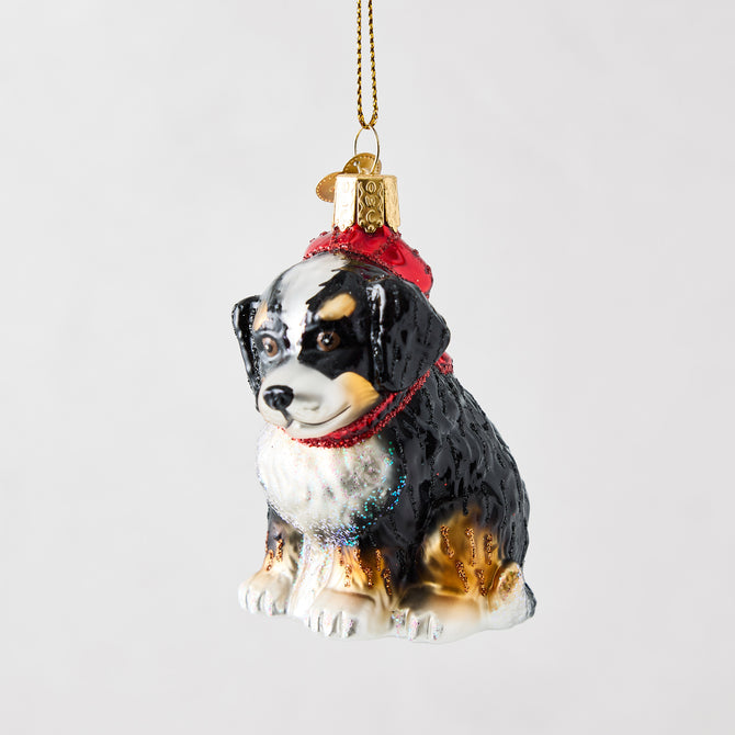 Hand-painted glass Bernadoodle ornament wearing a red hat with glitter accents and a gold hanging cord.