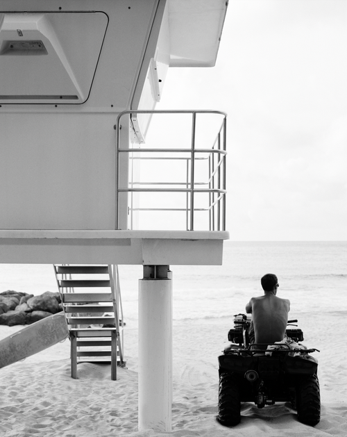 Black and white photograph by Matt Albiani of a lifeguard on an ATV facing the ocean next to a white lifeguard tower.