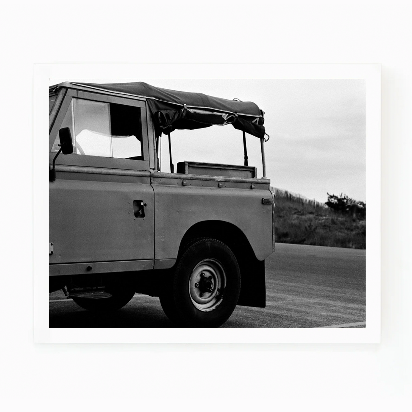 Black and white photograph of a vintage surf truck with a soft top, parked on a road near dunes.