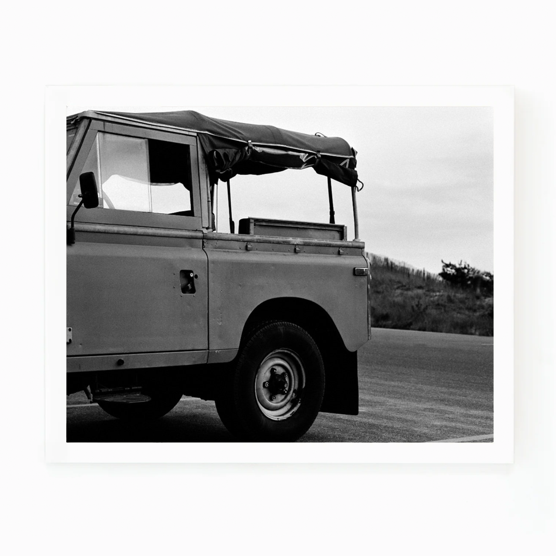 Black and white photograph of a vintage surf truck with a soft top, parked on a road near dunes.