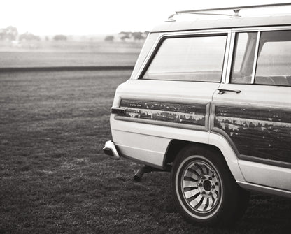 Black and white photograph of a vintage white Wagoneer with wood paneling parked in a grassy field.