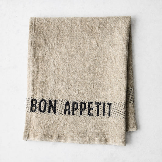 Camel khaki linen napkin with "BON APPETIT" in black lettering, folded on a white surface.