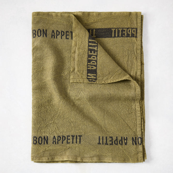 Folded green linen table runner with "BON APPETIT" in black text.