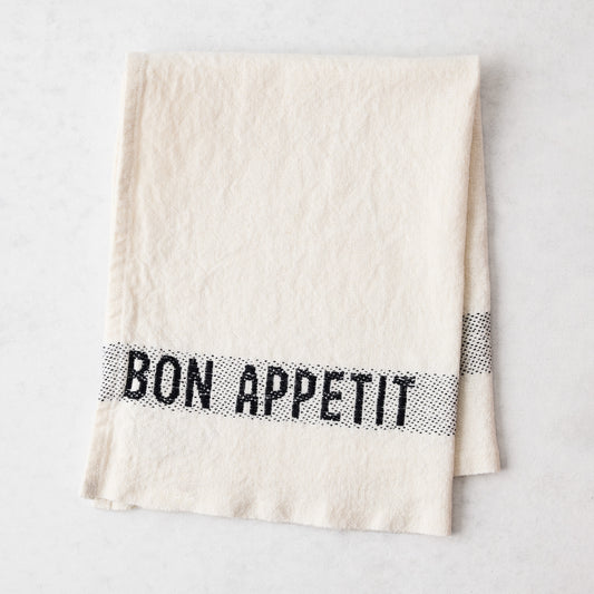 Off-white linen napkin with "BON APPETIT" in black lettering, folded on a white surface.