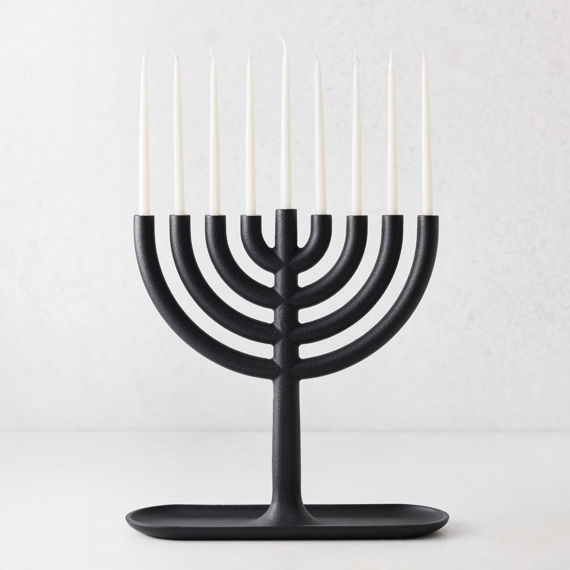 Modern black cast iron Hanukkah menorah by Josh Owen with a tray base and nine thin white taper candles.