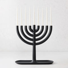 Modern black cast iron Hanukkah menorah by Josh Owen with a tray base and nine thin white taper candles.