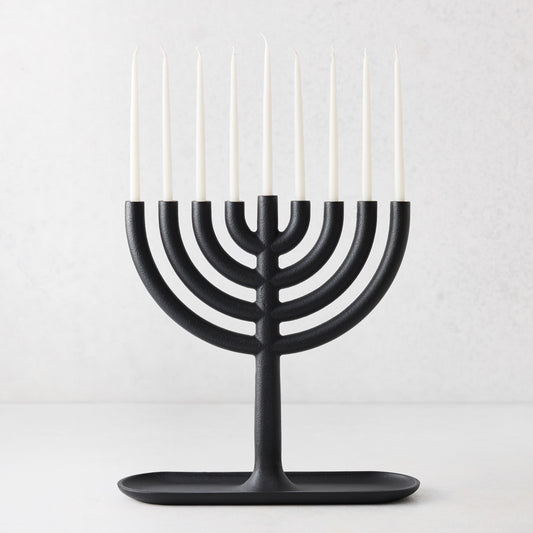 Modern black cast iron Hanukkah menorah by Josh Owen with a tray base and nine thin white taper candles.
