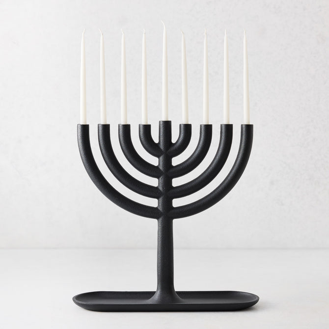 Modern black cast iron Hanukkah menorah by Josh Owen with a tray base and nine thin white taper candles.
