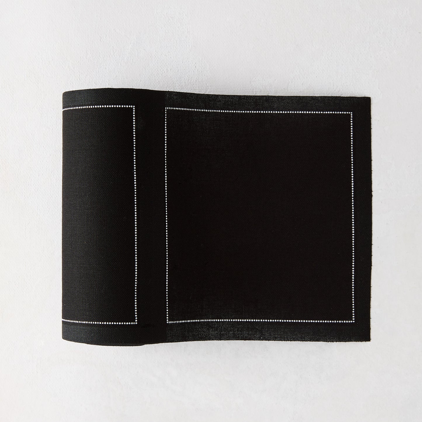 Black tear-off cotton cocktail napkin, partially unrolled, with white dotted border stitching.
