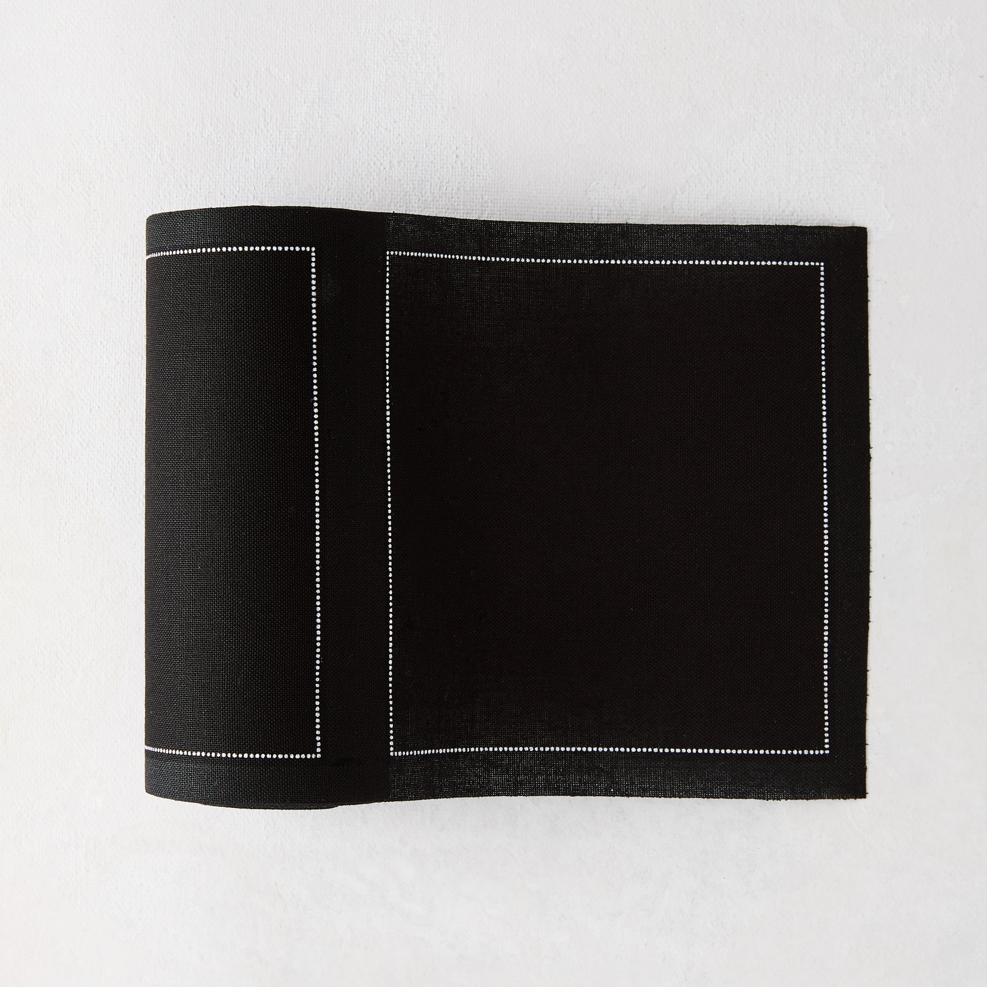 Black tear-off cotton cocktail napkin, partially unrolled, with white dotted border stitching.