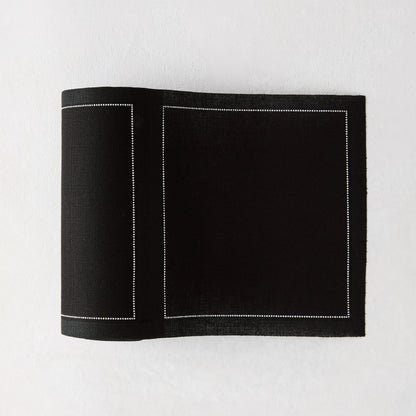 Black tear-off cotton cocktail napkin, partially unrolled, with white dotted border stitching.