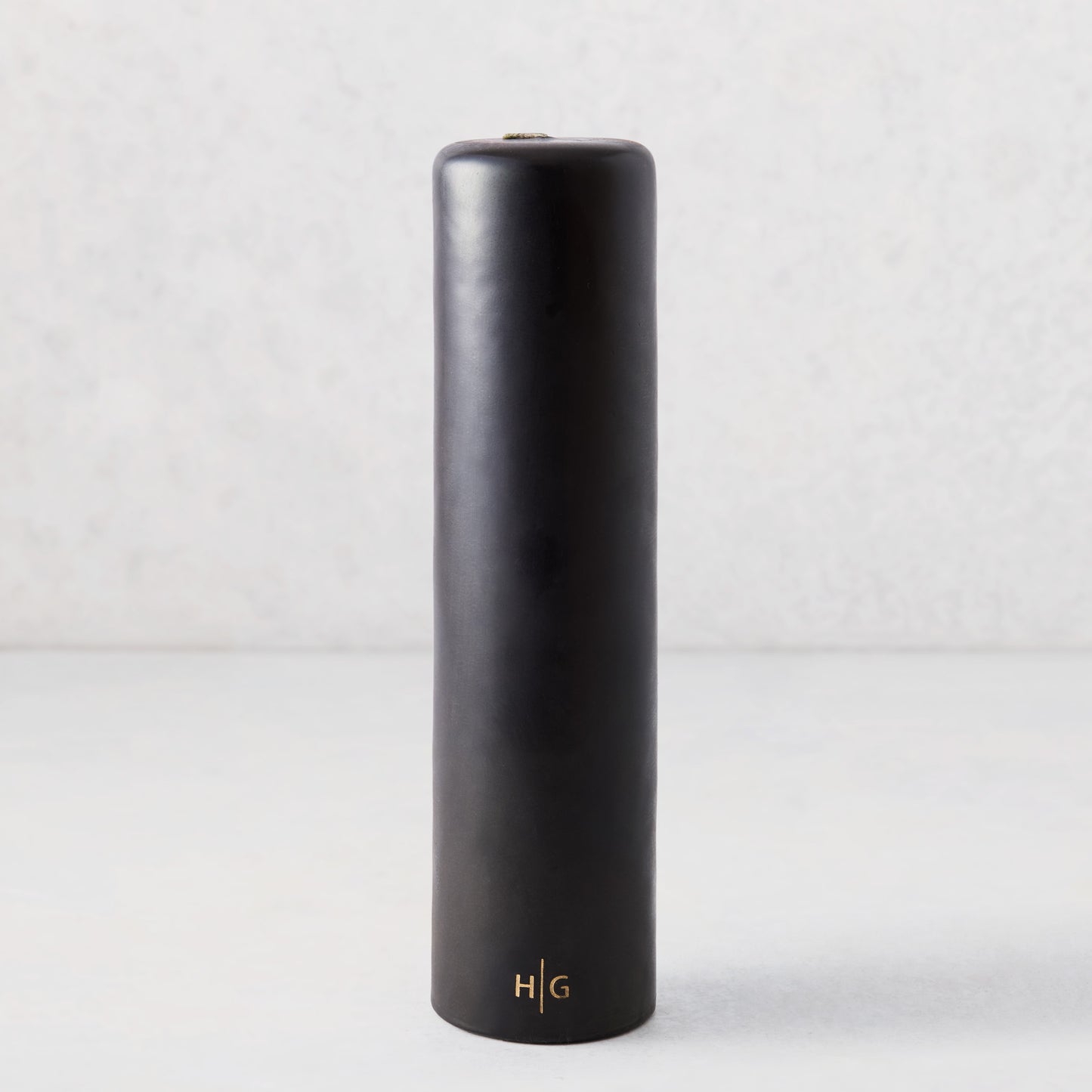 3" x 12" black unscented pillar candle with a gold embossed HG logo on a light grey background.