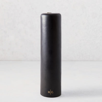 3" x 12" black unscented pillar candle with a gold embossed HG logo on a light grey background.