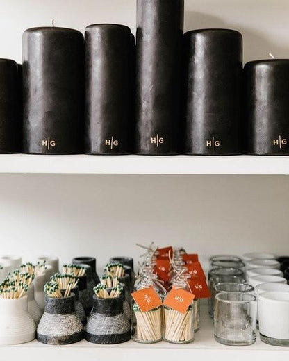 HG Black Unscented Pillar Candles, 3"x12", with gold HG logo, displayed on a shelf above matches and votives.