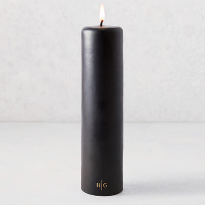 3x12 inch black unscented pillar candle with a gold HG logo and a lit cotton wick.