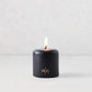 3x3 inch black hand-dipped pillar candle with a burning wick and gold embossed HG logo on a white surface.