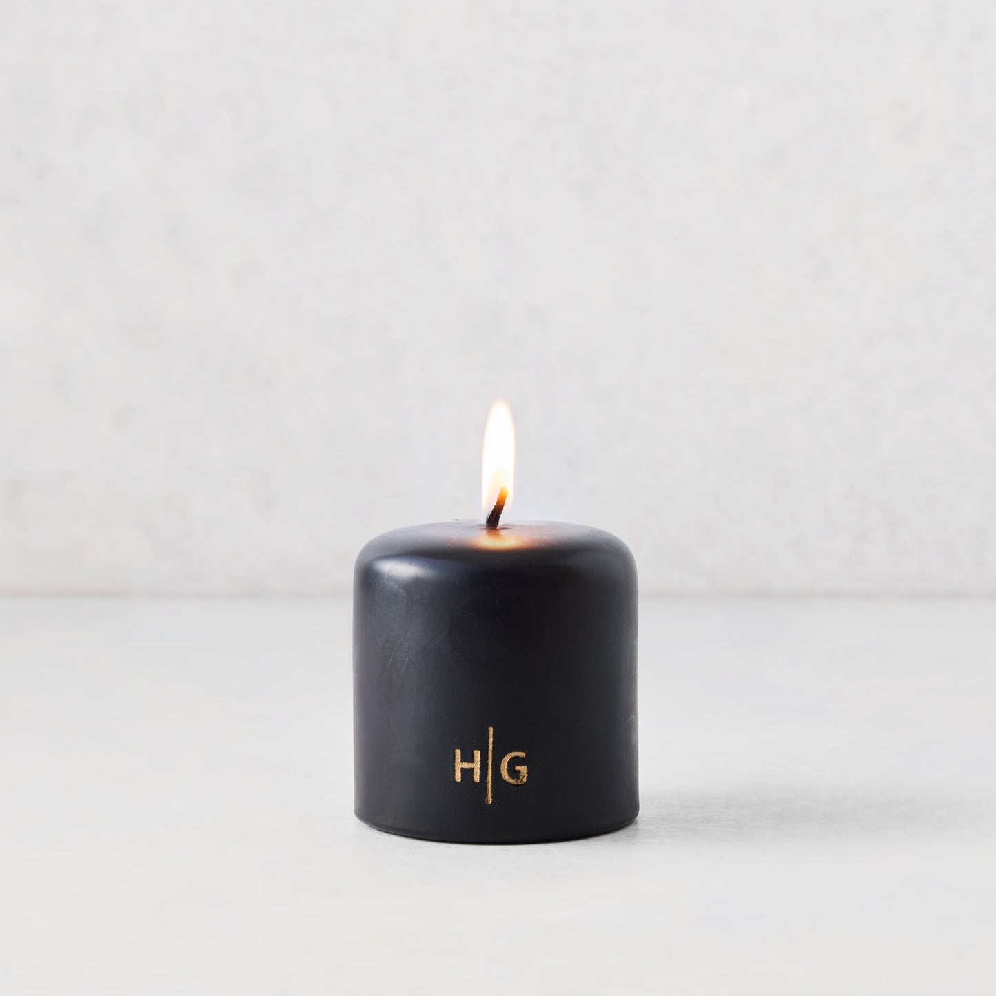 3x3 inch black hand-dipped pillar candle with a burning wick and gold embossed HG logo on a white surface.