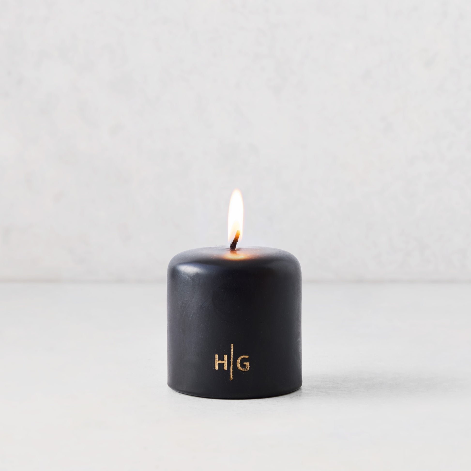3x3 inch black hand-dipped pillar candle with a burning wick and gold embossed HG logo on a white surface.